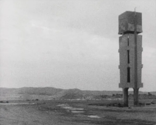A black and white photo, of a landscape with a mountain. On the right side there is a construction standing upon two columns, and a clear rectangular prism and a dark cube at the top, that resembles a robotic figure.